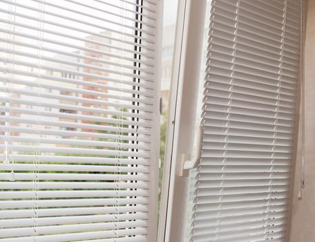 Fragment of the modern tilt and turn plastic window tilted inwards at the top with control handle and white horizontal window blinds 
