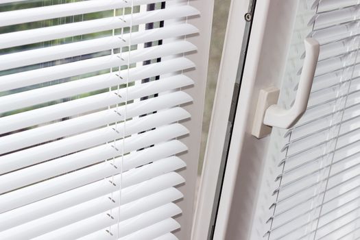 Fragment of the modern tilt and turn plastic window tilted inwards at the top with control handle and white Venetian blinds 
