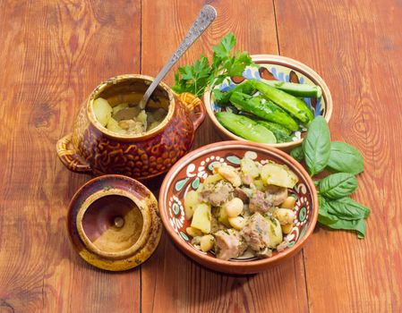 Ukrainian version of the dish Chanakhi in a clay bowl  - potatoes with meat, mushrooms and haricot beans roasted in a clay pot and lightly salted cucumbers on a surface of an old wooden planks
