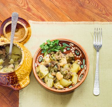 Top view of the Ukrainian version of the dish Chanakhi in a clay bowl and fork on a napkin - potatoes with meat, mushrooms and haricot beans baked in a clay pot on wooden surface
