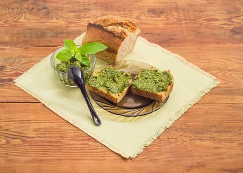 Two sandwiches with sauce pesto, sauce pesto in small glass bowl with small black spoon and decorated with green basil twig, part of bread on a napkin on a surface of  old wooden planks
