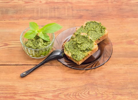 Two sandwiches with pesto on glass saucer and small black spoon, sauce pesto in small glass bowl decorated with green basil twig on a surface of old wooden planks
