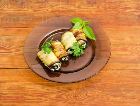 Eggplant rolls with tuna and processed cheese filling decorated with parsley and basil twigs on a dark glass dish on a surface of an old wooden planks
