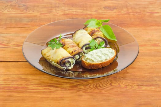 Eggplant rolls with tuna and processed cheese filling decorated with parsley and basil twigs and sandwich with stuffing on a dark glass dish on a surface of an old wooden planks

