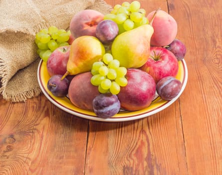 Several apples, pears, plums, peaches and clusters of white grapes on the big yellow dish on a surface of old wooden planks with sackcloth
