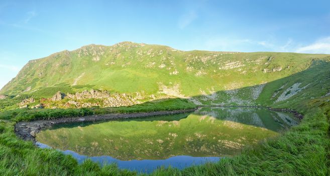 Panorama of the mountain lake located in an open circus with mountain slope reflected in a water in summer morning

