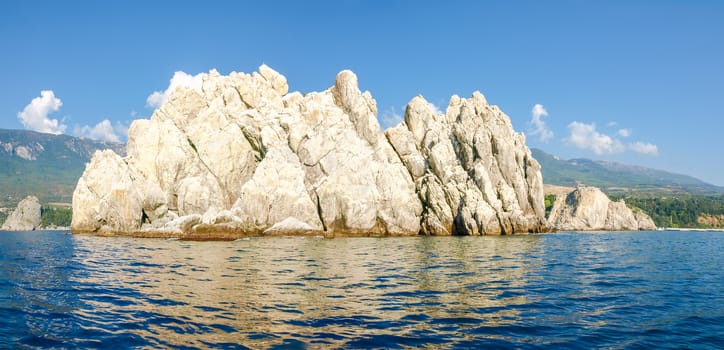 Panorama of the single limestone rock in the sea on a background of the sky and shore with rocks and mountain slopes overgrown with forest
