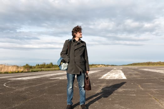 Young man in blue jeans and grey coat standing on the runway going on vacation on air plane with brown leather bag in hand. White aircraft on the background.