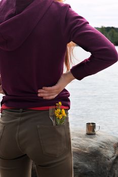 Handsome female tourist wearing brown jeans and purple sweatshirt with fork and yellow flowers in her pocket stands on river bank and enjoys view of mountains in summer. Hike or trip concept. Vertical.
