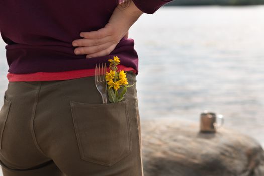 Handsome female tourist wearing brown jeans and purple sweatshirt with fork and yellow flowers in her pocket stands on river bank and enjoys view of mountains in summer. Hike or trip concept. Horizontal