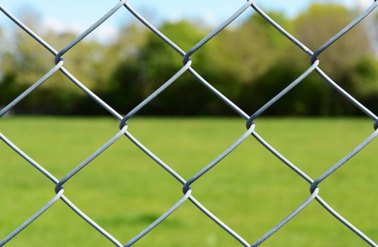 Metal chain link fencing in selective focus. Beyond the secure fence lies an empty green field, lined by trees.