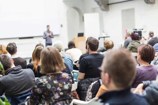 Male speaker giving presentation in lecture hall at university workshop. Audience in conference hall. Rear view of unrecognized participant in audience. Scientific conference event.