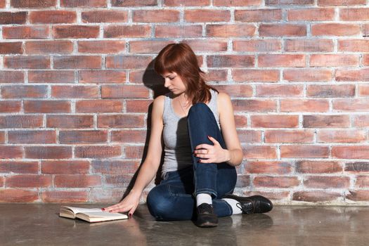 Beautiful young red hair woman in jeans with book sitting on floor near brick wall. Attentively reading an interesting book