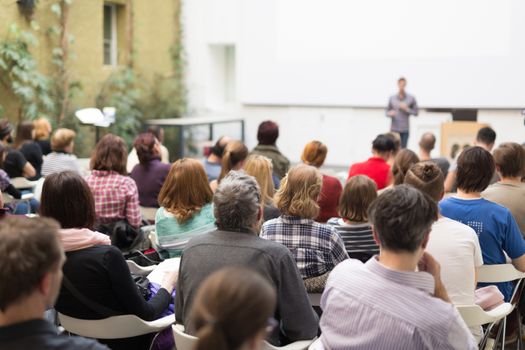 Male speaker giving presentation in lecture hall at university workshop. Audience in conference hall. Rear view of unrecognized participant in audience. Scientific conference event.