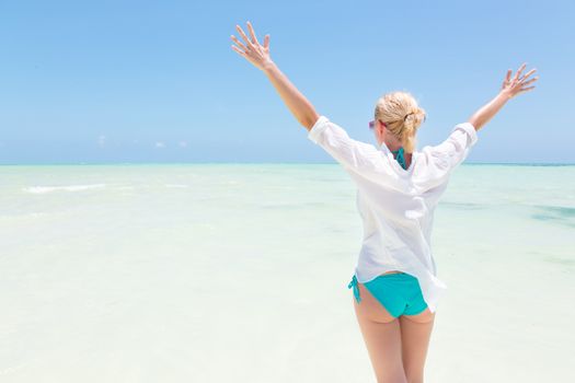 Happy woman, arms rised, enjoying, relaxing joyfully in summer at white tropical beach. Beautiful caucasian model wearing white beach tunic on vacations on Paje beach, Zanzibar, Tanzania.