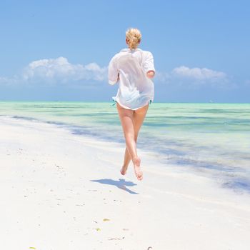 Happy woman having fun, enjoying summer, running joyfully on tropical beach. Beautiful caucasian model wearing white beach tunic on vacations on picture perfect Paje beach, Zanzibar, Tanzania.