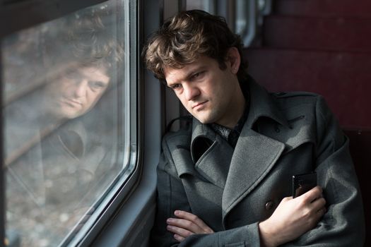Traveling by train. Sad man traveling by train, looking through the window and thinking about unrequited love squeezing the phone in his hand. alone in an empty train wagon. Real people series. Toned picture.
