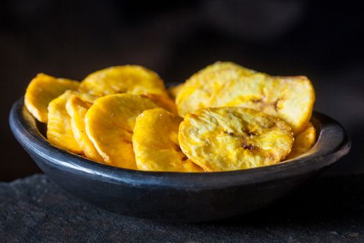 Plantain fried coins in a traditional black clay bowl on black background