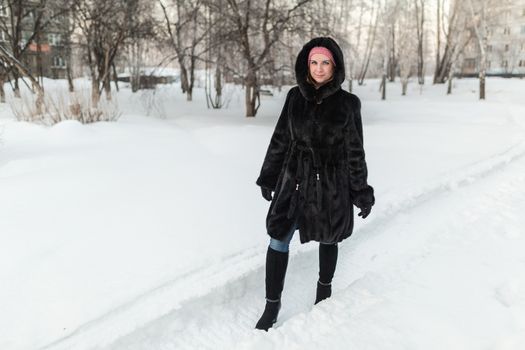 the girl in a black fur coat goes on a snow-covered footpath