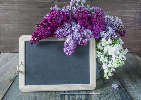 Blank chalkboard and lush multicolored bouquet of lilac flowers in a metal pitcher on an old wooden background