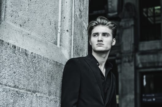 Handsome blond young man, blue eyes, leaning against white, rough stone wall, looking up to a side