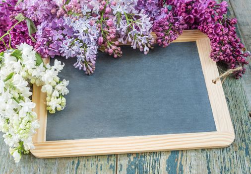 Flower border composed of lush multicolored bunches of lilac flowers around of empty black chalkboard on the old wooden background