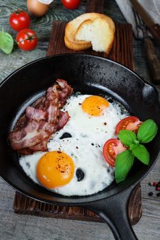 Breakfast in a rustic style: two fried eggs, strips of bacon, fresh tomatoes and basil in a cast iron skillet and toasts on an old wooden table 