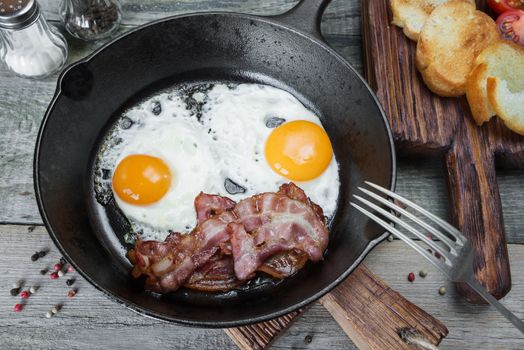 Breakfast in a rustic style: two fried eggs, strips of bacon  in a cast iron skillet and toasts on an old wooden table 