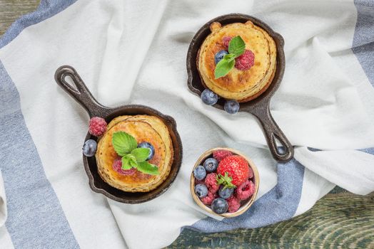 Pancakes in cast-iron frying pans with fresh berries on the background of the old wooden boards covered with a linen tablecloth