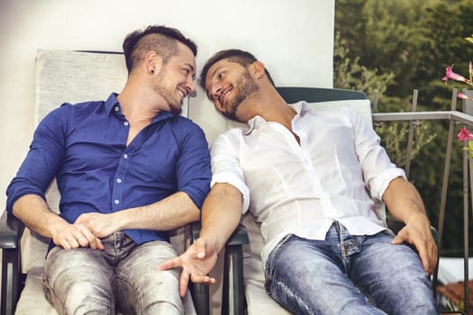 Two gays sitting on chairs at balcony holding hands and looking at each other.