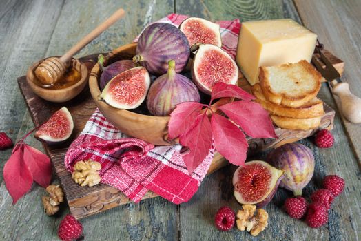 Beautiful autumn still life: ripe figs in a wooden bowl, red raspberry, toasts, cheese, honey and a checkered napkin on old cutting board as well as autumn leaves on the old wooden table