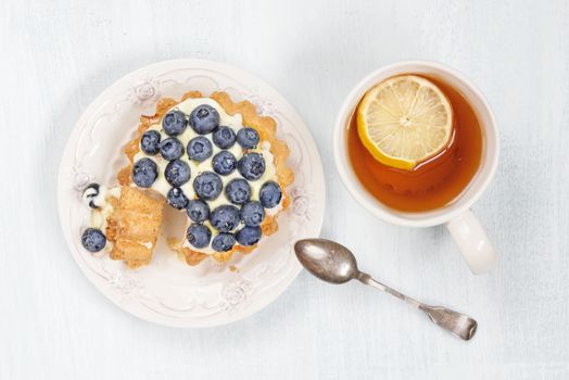 Sweet cake with blueberries, butter cream and syrup, as well as a cup of black tea with a slice of lemon, top view