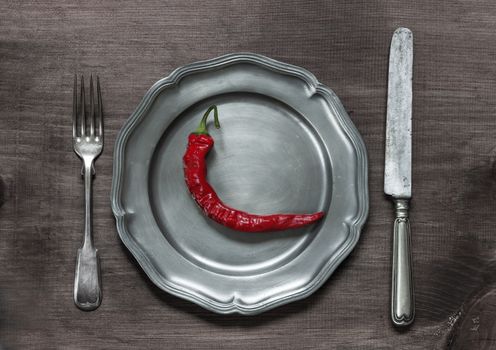 Red hot chilli pepper on vintage pewter plate, as well as fork and knife on old brown dark wooden board