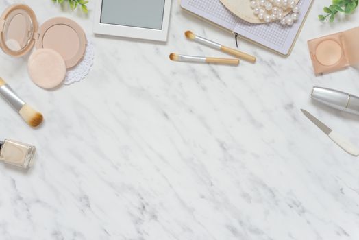Feminine workspace on table desk with white smartphone, lipstick; pearl necklace, beige compact powder, cosmetic brushes and pink nail polish on marble stone background; top view, flat lay