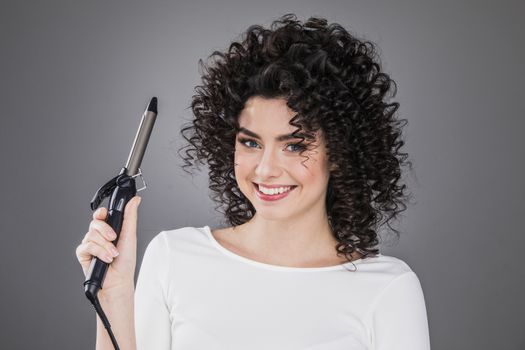 Portrait of beautiful smiling woman with curly hair holding curling iron, gray background