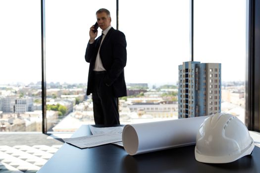 Businessman in construction company office, model of modern multi storey residential building house, hardhat, blueprint and documents on table