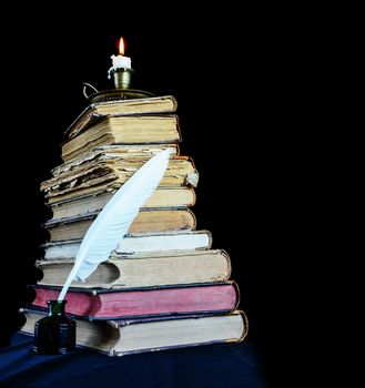 High stack of old books, a burning candle in a candlestick and a white feather in the inkwell