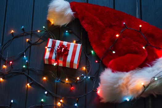 Santa claus hat, fir tree branch with decoration, gift boxes With bright bulbs flashlights garland on a black background New year and Christmas background