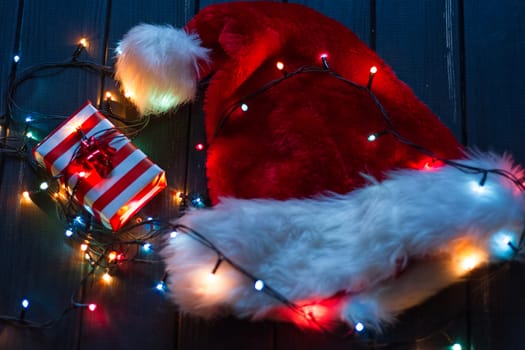 Santa claus hat, fir tree branch with decoration, gift boxes With bright bulbs flashlights garland on a black background New year and Christmas background