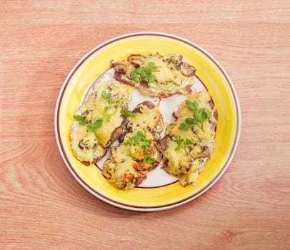 Top view of several pork chops baked with onion, mushrooms and cheese and decorated with twigs of the parsley on the yellow and white dish on a wooden surface
