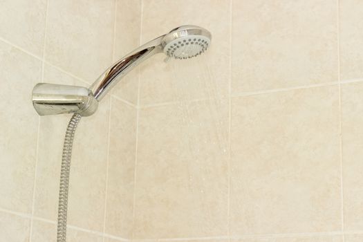 Water flowing from shower head with turn lever of a spray settings and metal shower hose in holder on a background of a wall with beige tiles
