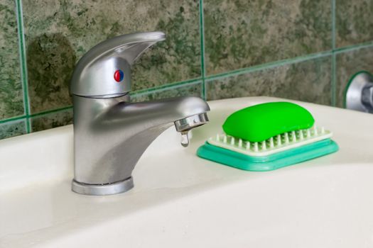 Single handle mixer tap mounted on a wash basin with drop of water on background of a wall with green tiles
