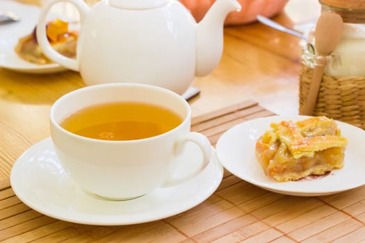White cup with a herbal tea and a piece of cake on a saucer on the bamboo napkin on a blurred background of a teapot and other tea appliances
