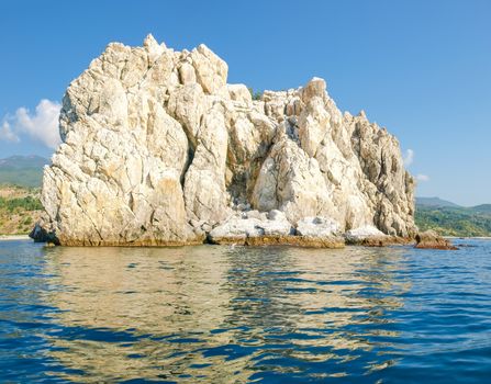 Single limestone rock in the sea on the background of the shore with the mountain slopes and the sky
