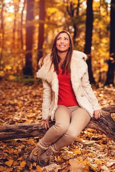 Beautiful smiling girl enjoying in forest in autumn. She is sitting on the tree and thinking.