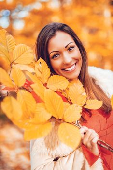 Portrait of a cute smiling young woman enjoying in forest in autumn. She is holding yellow leaves and looking at camera.