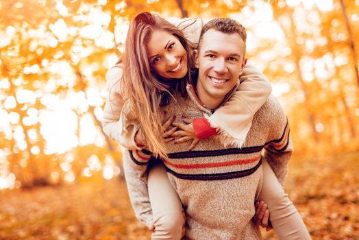 Portrait of a young smiling couple enjoying a piggyback in sunny forest in autumn colors and looking at camera.