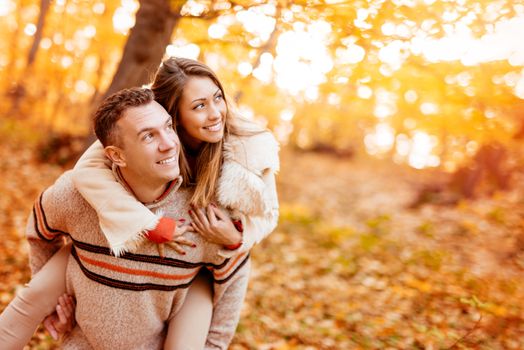 Beautiful young couple enjoying a piggyback in sunny forest in autumn colors.