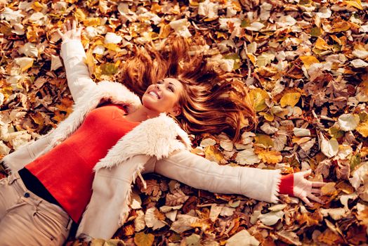 Cute smiling young woman lying on the falls leaves and enjoying in the nature in autumn.