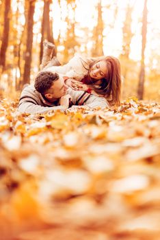 Beautiful smiling couple enjoying in sunny forest in autumn colors. They are lying on the falls leaves and having fun.